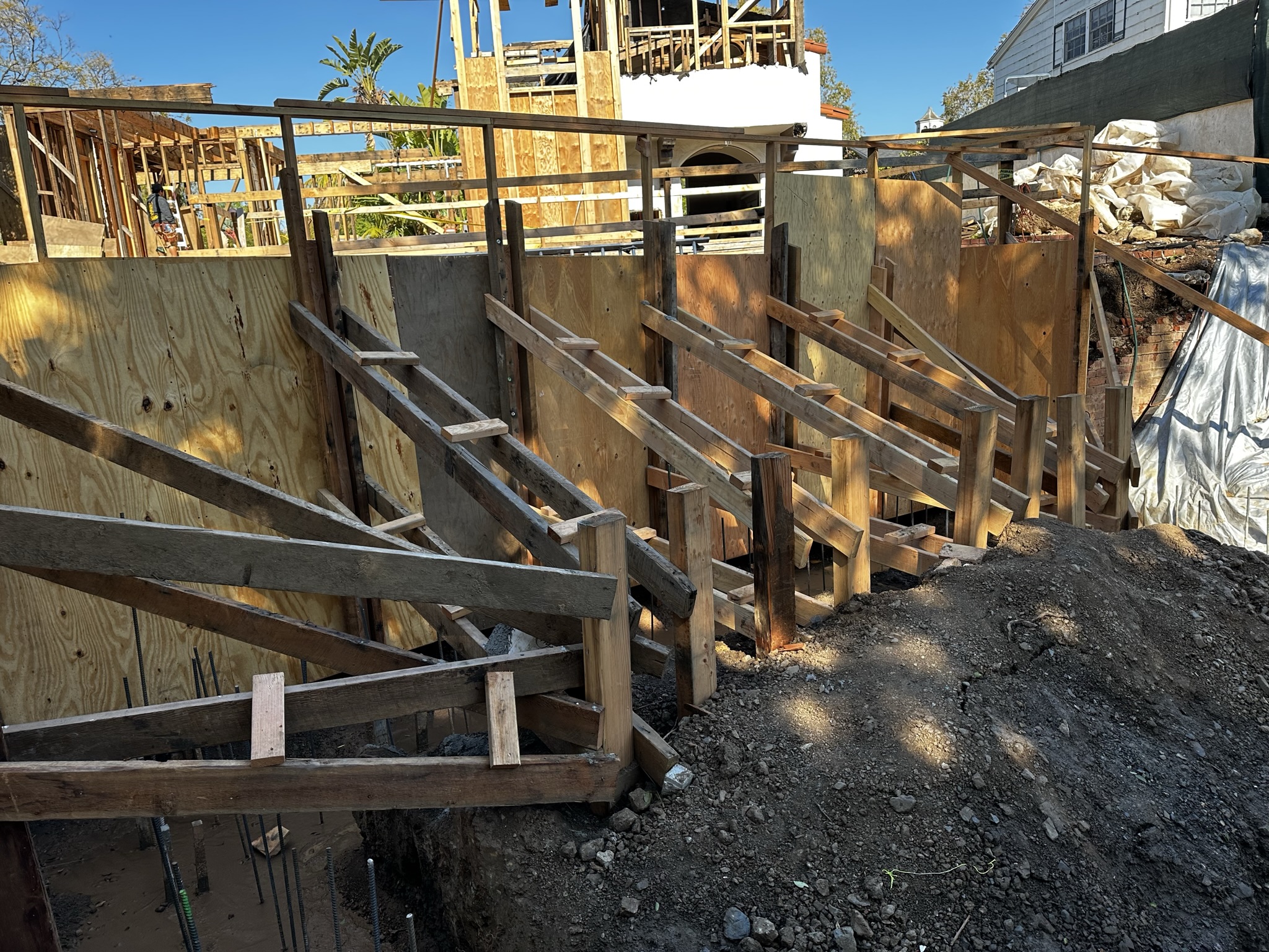 A construction site showing wooden shoring and bracing structures supporting excavation walls, with framing of a house visible in the background.