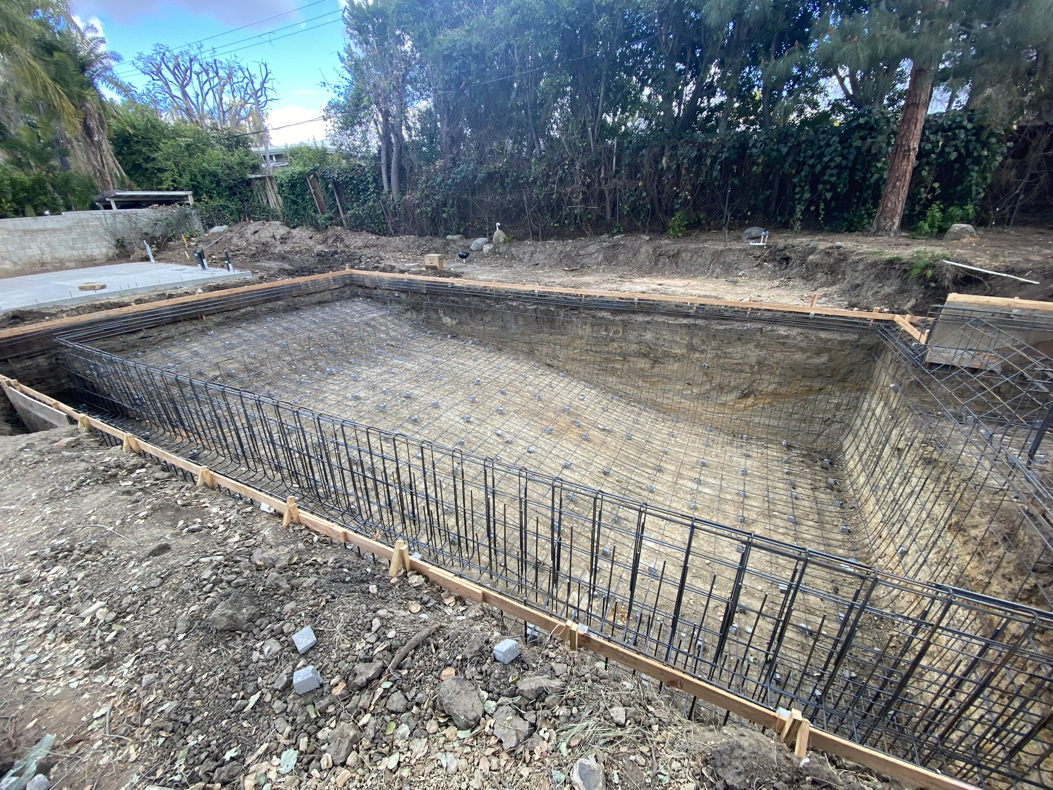 A large excavation site for a swimming pool featuring detailed rebar grid reinforcement and wooden formwork for structural support.