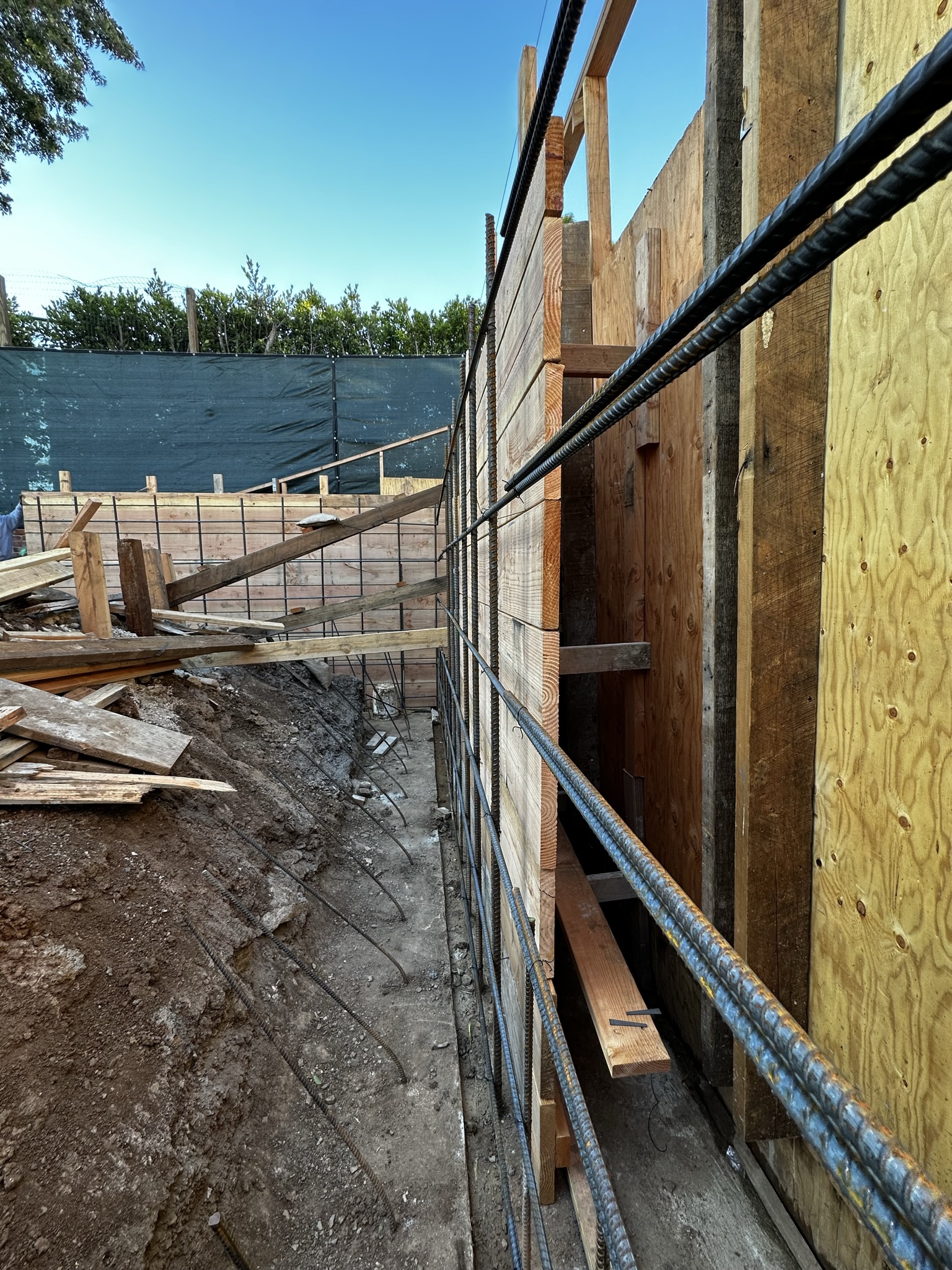 Close-up view of a construction site showing rebar reinforcement along a plywood formwork, prepared for pouring a concrete retaining wall with a clear blue sky in the background.